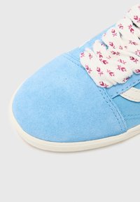 Gros plan sur le bout d'une basket en daim bleu clair avec une semelle blanche et des lacets blancs épais ornés de petits motifs floraux rouges.