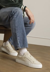 Beige suede sneakers with a white rubber sole, intricate stitching, and matching laces, paired with light blue jeans and white socks.