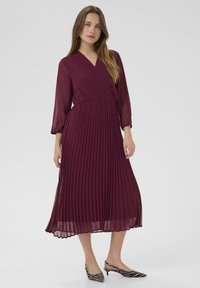Rochie burgundy plisată cu un decolteu înfășurat și mâneci lungi din tul. Asortată cu pantofi cu vârf ascuțit cu un model de zebra alb-negru.