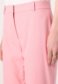 Różowe spodnie o gładkiej fakturze, z płaskim przodem, kieszeniami bocznymi i dopasowanym krojem. Ręka spoczywa na udzie, nosząc pierścionek.
