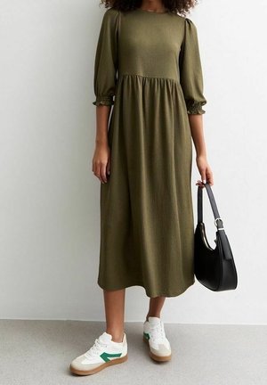 Robe vert olive, mi-longue, avec des manches bouffantes et des poignets élastiques, associée à des baskets blanches avec des accents verts et un sac à main noir.