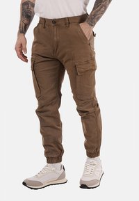 Pantaloni cargo in tessuto marrone, con molteplici tasche, polsini a tubo e una vestibilità comoda. Abbinati a scarpe da ginnastica chiare.