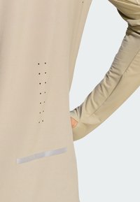 Béžový dlhý rukáv s malými ventilačnými otvormi a reflexným pruhom na dolnej časti chrbta, ruka zasunutá do bočného vrecka.