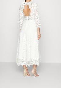 Valkoinen pitsimekko, jossa on pitkät hihat, korkea kaula-aukko, avoin selkä ja aaltoileva helma. Yhdistettynä nude-värisiin korkokenkiin.