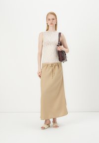 Hedvábný top bez rukávů v off-white barvě s kulatým výstřihem, zkombinovaný s béžovou maxi sukní na stahování. Modelka drží hnědou koženou tašku.