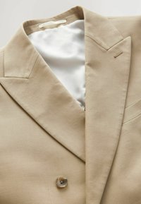 Blazer beige con colletto strutturato, chiusura a un bottone e rivestimento bianco liscio. Presenta cuciture minimaliste e un sottile dettaglio di metallo.