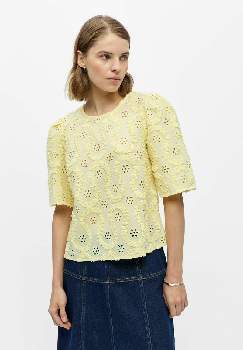 Femme aux cheveux courts portant un chemisier brodé jaune pâle avec des manches bouffantes et une jupe en jean bleu foncé, debout sur un fond blanc.