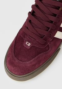 Nahaufnahme eines burgunderfarbenen Wildledersneakers mit dicken passenden Schnürsenkeln, cremefarbenen Seitenstreifen und einer strukturierten braunen Gummisohle.