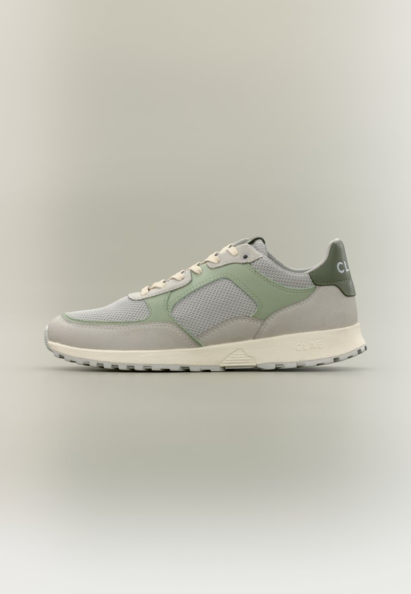 Αθλητικό παπούτσι σε ανοιχτό γκρι και πράσινο χρώμα με πάνελ από πλέγμα, μπεζ κορδόνια και λευκή σόλα, εμφανίζεται από το πλάι σε απλό ουδέτερο φόντο.