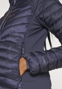 Marineblauwe pufferjack met een gestikt patroon, voorzien van een zijritszak en elastische manchetten. De stof is lichtgewicht en waterafstotend.