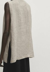 Helle beige ärmellose Bluse aus strukturiertem Stoff, mit seitlichen Schlitzen und lockerer Passform. Der Rücken ist etwas länger als die Vorderseite.