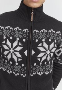 Schwarzer gestrickter Reißverschluss-Pullover mit hohem Kragen, der komplizierte weiße und graue Schneeflockenmuster sowie ein bronzefarbenes Reißverschlussdetail aufweist.