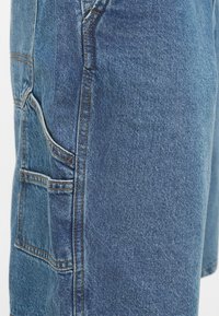 Primo piano di tessuto denim blu con più tasche cucite e cuciture visibili su un paio di jeans o pantaloncini.