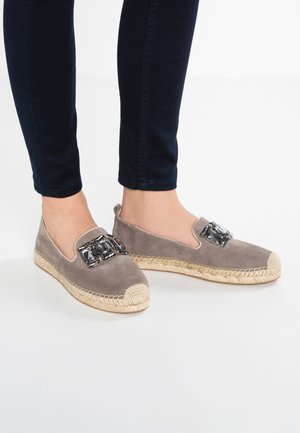 Fødder iført taupe ruskind espadriller med flettede såler og store rektangulære klare ædelstensdekorationer, kombineret med mørke, stramme jeans.