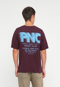 Maroon cotton t-shirt featuring a large blue graphic print on the back that includes "PNC PADEL NEW YORK SPORT CLUB" and additional text below.