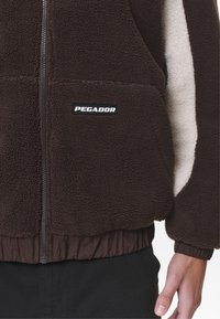 Giacca in pile marrone con zip frontale, caratterizzata da un interno crema e etichetta PEGA-DOR sulla tasca. Polsini e orlo elastici. Superficie testurizzata.