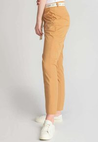Pantaloni beige con vestibilità slim, realizzati in tessuto liscio. Presentano un cinturino bianco con testo stampato, tasche laterali e dettagli minimi.