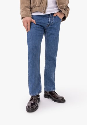 Mężczyzna ubrany w niebieskie jeansy o prostych nogawkach, ciemnobrązowe skórzane buty, beżową kurtkę i białą koszulę, z jedną ręką w kieszeni.