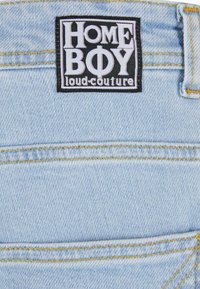 Lys blå denimstoff med synlige sømmer. Har et svart merke med hvit tekst som leser "HOME BOY loud-couture" festet på livet.