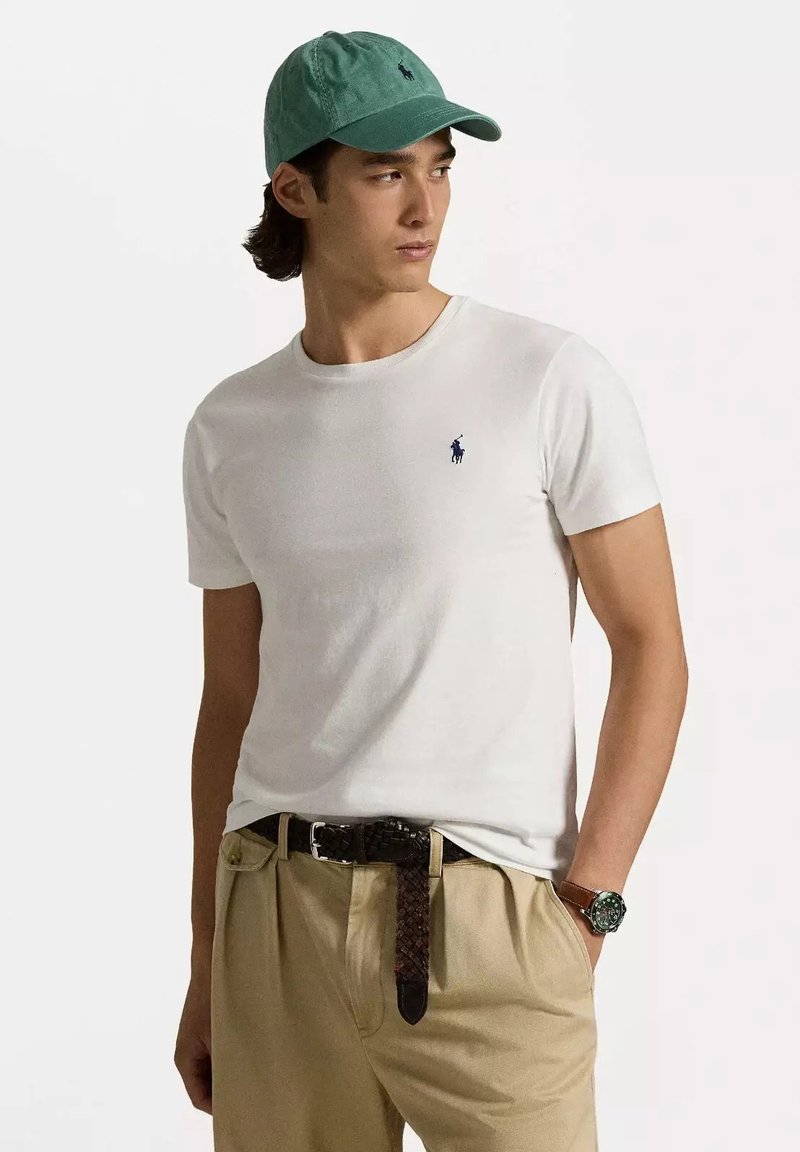 T-shirt bianco a maniche corte con un piccolo logo blu, abbinato a pantaloni khaki e una cintura intrecciata marrone per aggiungere texture. Cappellino teal in testa.