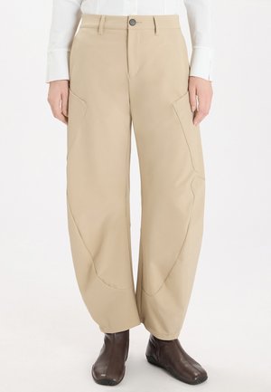Pantaloni beige a gamba larga con tasche laterali indossati con una camicia bianca e stivaletti marrone scuro, mostrati dalla vita ai piedi.
