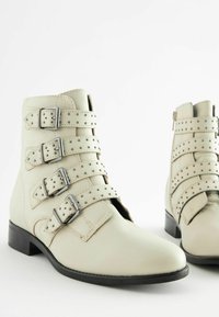 Beige leather ankle boots featuring four adjustable buckle straps with studs, and a low black heel. Smooth texture with rounded toe design.