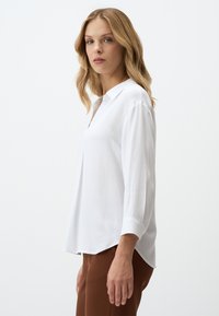 Chemise blanche à manches longues avec un col et un motif texturé subtil. L'ourlet est légèrement plus long à l'arrière, associée à un pantalon marron.