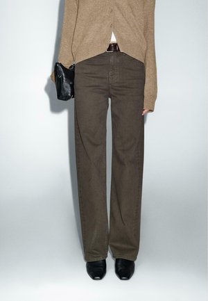 Osoba nosząca brązowe szerokie spodnie z wysokim stanem, beżowy kardigan, czarne buty i trzymająca małą czarną torebkę na jednolitym tle.