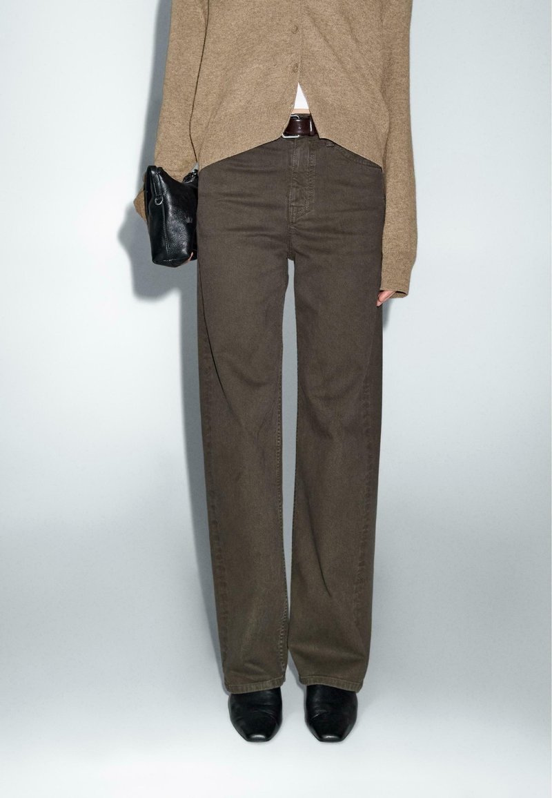 Personne portant un pantalon marron taille haute à jambes larges, un cardigan beige, des chaussures noires et tenant un petit sac à main noir sur un fond uni.