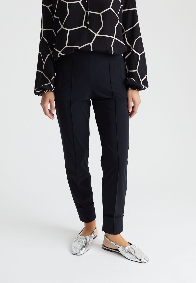 Zwarte tapered broek met plooien, gecombineerd met een blouse met lange mouwen en een patroon. Zilveren instapschoenen met een gestructureerde afwerking.