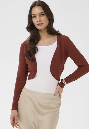 Mujer de cabello largo y oscuro con una blusa blanca, cárdigan corto color óxido, falda beige y pulsera plateada, posando con una mano en la cadera.