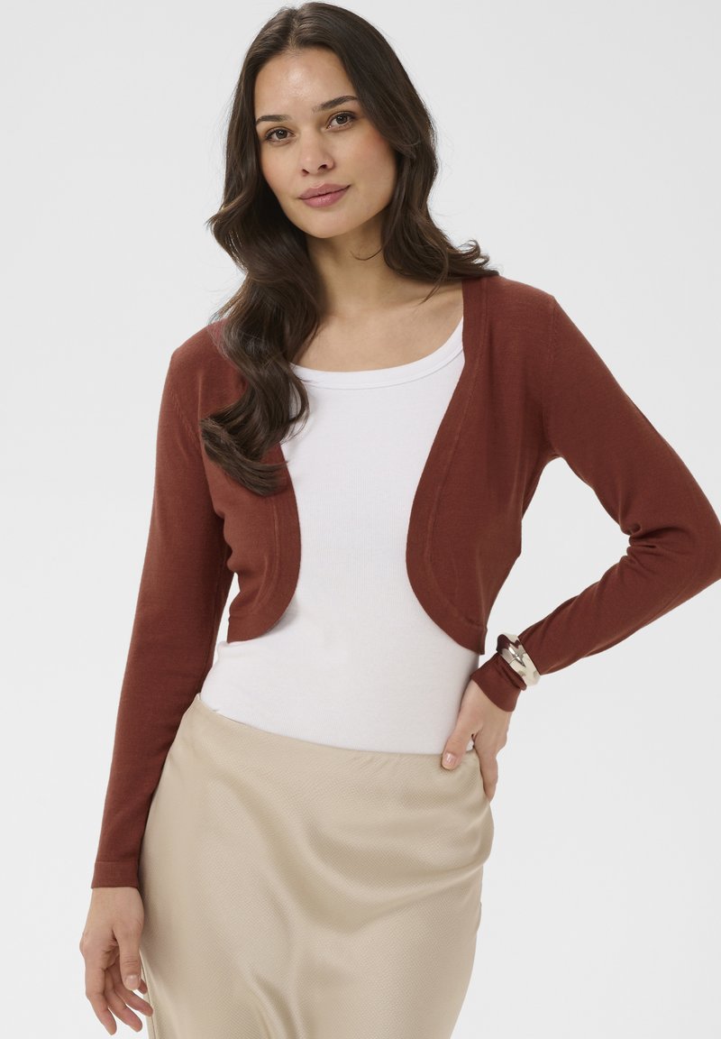 Femme aux longs cheveux noirs portant un haut blanc, un cardigan court couleur rouille, une jupe beige et un bracelet argenté, posant avec une main sur la hanche.