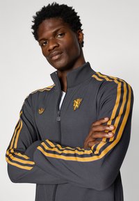 Σκούρο γκρι ζακέτα φόρμας με φερμουάρ και κίτρινες και μωβ ρίγες στα μανίκια. Διαθέτει κεντημένο λογότυπο στο στήθος και υφή υφάσματος.