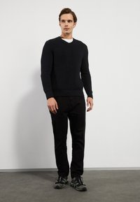 Marineblauer gerippter V-Ausschnitt-Pullover mit langen Ärmeln, kombiniert mit schwarzen Hosen und Sportschuhen, vor einem neutralen Hintergrund.