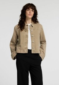 Beige denimjacka med krage, framknäppning med tryckknappar och lång ärm. Bärs över en vit topp och svarta byxor.