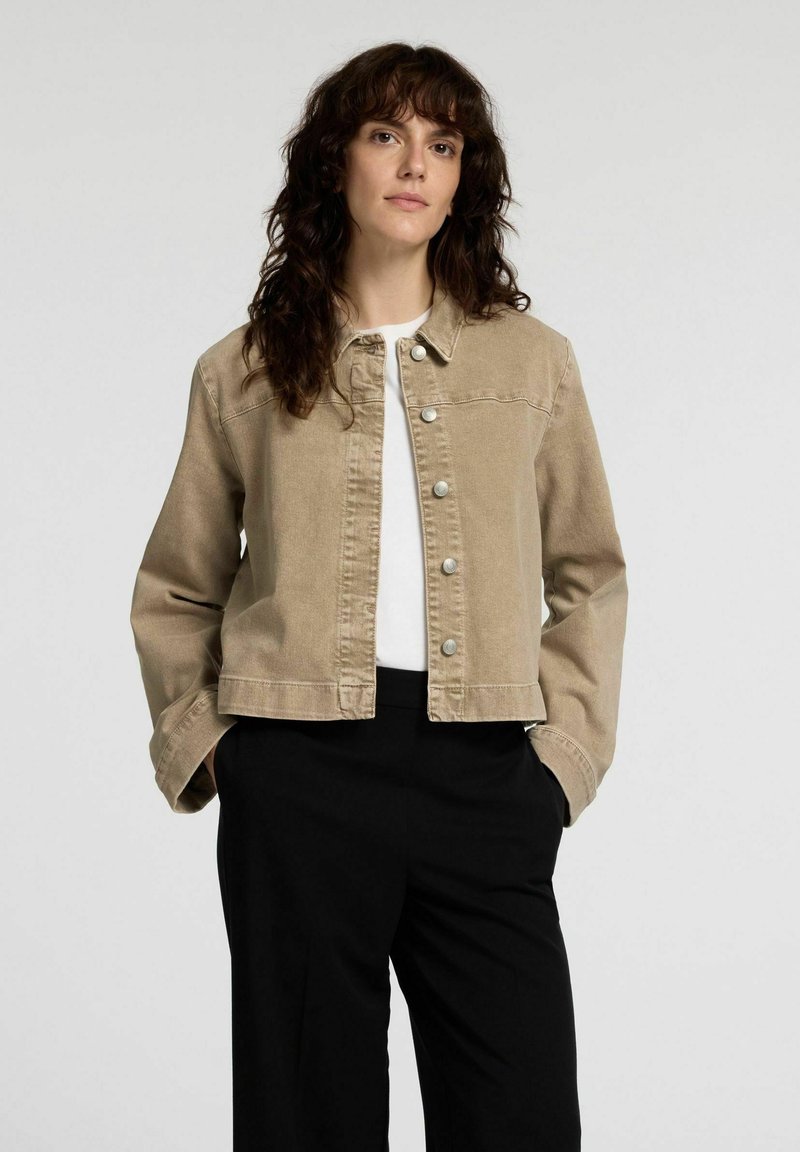 Beige denimjacka med krage, framknäppning med tryckknappar och lång ärm. Bärs över en vit topp och svarta byxor.