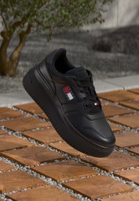 Black leather sneaker with a textured sole, featuring a colorful logo patch and tonal laces, set against a brick and gravel surface.