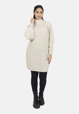 Donna che indossa un abito maglia beige a coste, leggings neri e stivali militari neri con lacci, posando con una mano vicino al colletto.