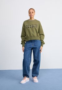 Maslinasto zelena Nike dukserica s velikim otisnutim logotipom, u kombinaciji s labavim plavim trapericama i svijetloružičastim tenisicama s bijelim potplatom.