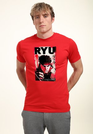 Ung mand iført rød t-shirt med grafik af Ryu fra Street Fighter, stående med hænderne bag ryggen mod en ensfarvet baggrund.