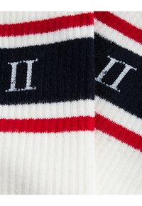 Weiße Socken mit roten und marineblauen Streifen, mit geripptem Textil. Die Zahl "II" ist in Hellgrau auf einem dunklen Hintergrund gestickt.