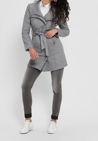 Cappotto in misto lana grigio con vita con cintura, colletto a revers e dettagli con tasche con zip, abbinato a pantaloni grigi slim-fit e sneakers bianche.
