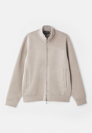 Chaqueta cremosa con cremallera, de tela texturada, con cuello alzado y dos bolsillos frontales. Presenta un patrón sutil y puños de punto.