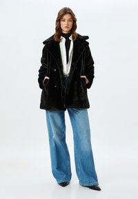 Sort faux fur dobbeltrækket frakke med store klapper, sidelommer, båret over en hvid skjorte og brede blå denim jeans.