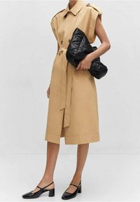 Beżowy płaszcz trench z przesadnie dużymi rękawami, podwójnym zapięciem na guziki, wiązanym w talii i w bocznym rozcięciu. Czarna pikowana torebka kopertówka i czarne buty na słupku.