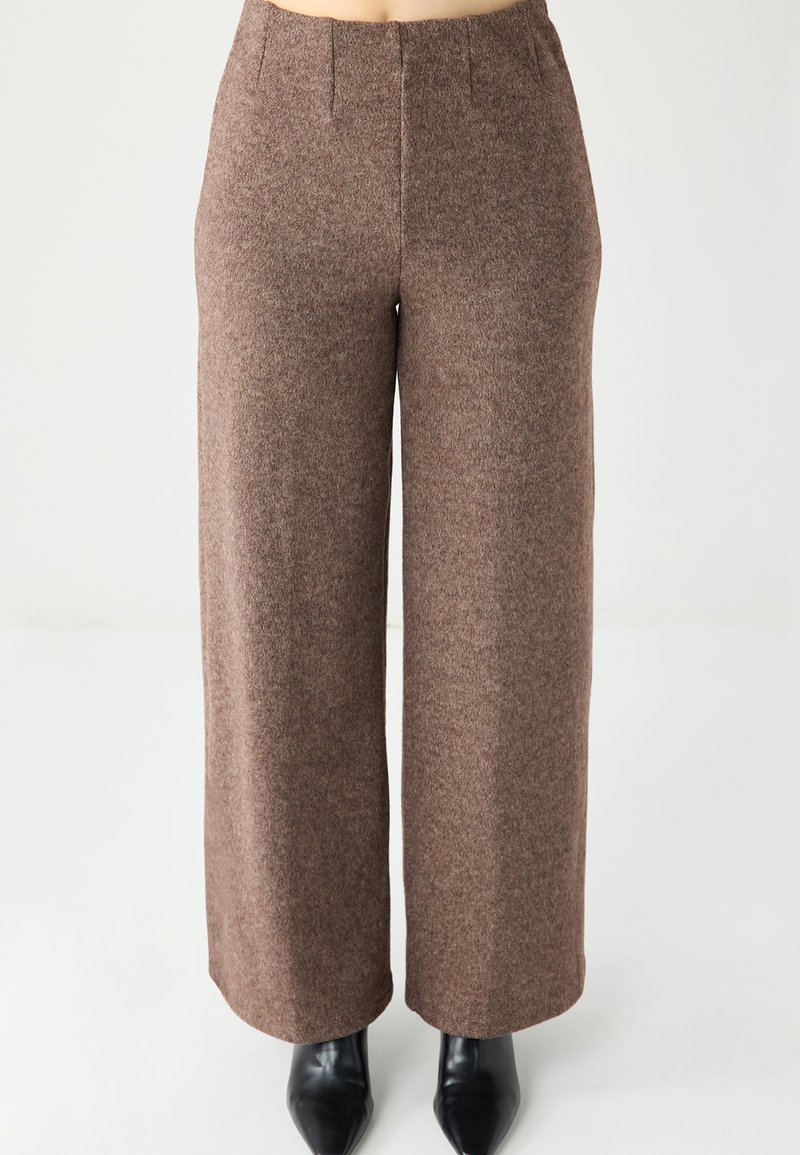 Pantalon marron à jambes larges en tissu texturé, avec des plis devant et une taille haute. Porté avec des bottes noires à bout pointu.