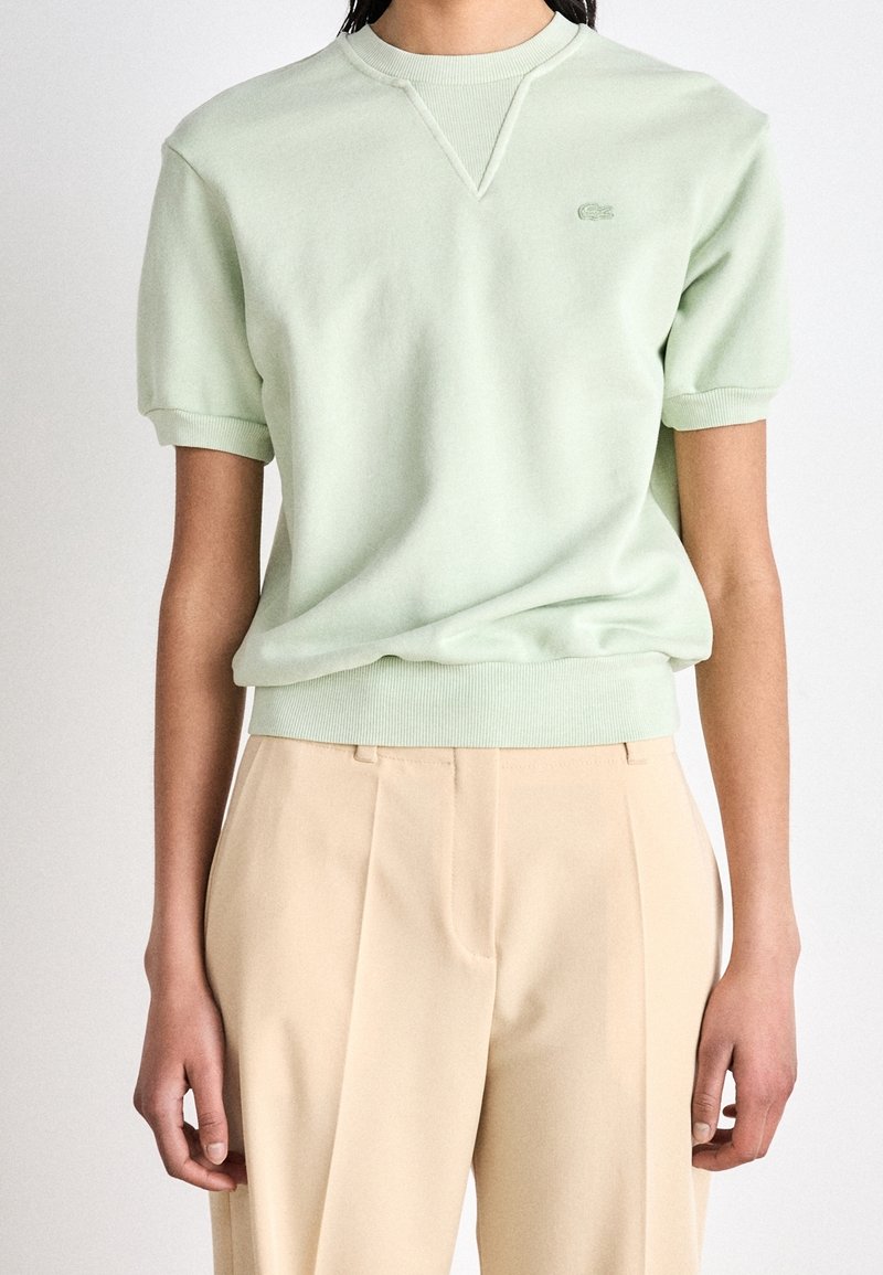 Light green cotton-blend short-sleeve sweatshirt with ribbed neckline and cuffs. Features a subtle logo on the chest. Paired with beige trousers.