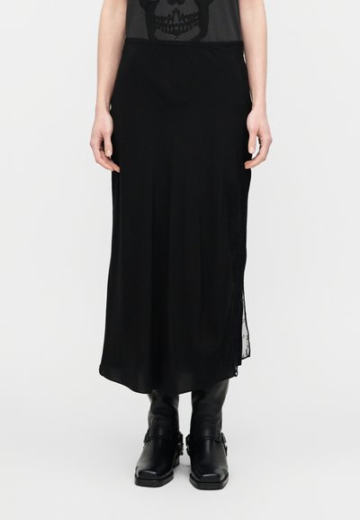 Jupe longue noire en matériau léger, avec un design fluide, comportant une fente latérale et une bordure en dentelle. Associée à des bottines noires.