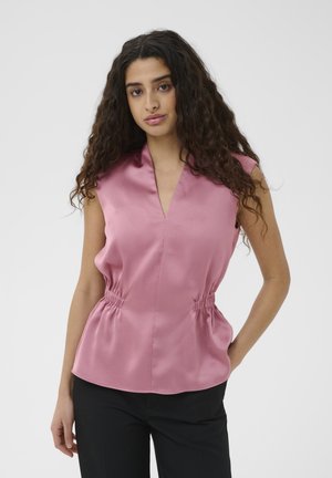 Naine pikkade lokkis juustega, kellel on seljas varrukateta roosa pluus V-kaelusega ja mustad püksid, seisab lihtsa tausta ees.