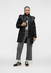 Ung kvinde iført en sort pufferjakke, grå cargo-bukser, sorte sneakers og en argyle sweater, stående foran en ensfarvet hvid baggrund.